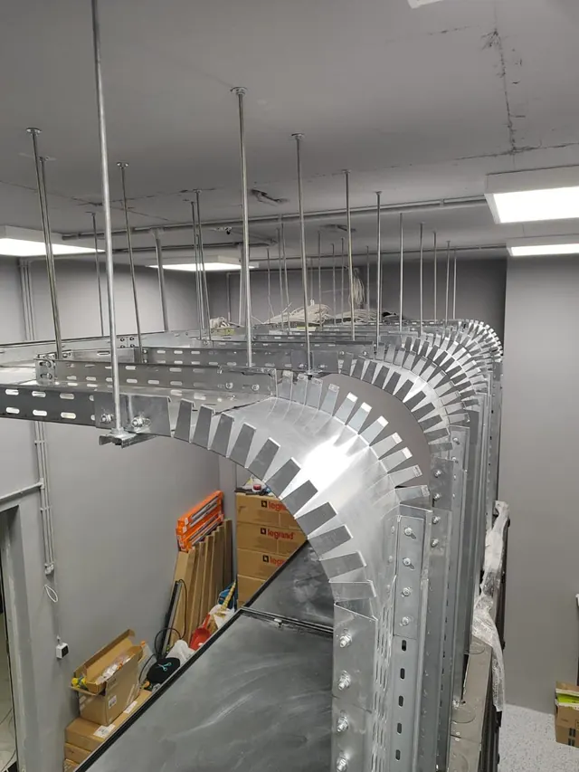 Overhead galvanised cable tray turning a wide radius corner through a finished office ceiling void, suspended on threaded rod