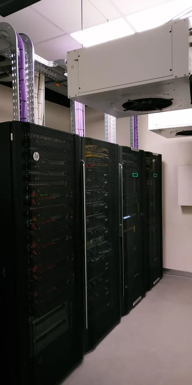 A row of installed and powered equipment cabinets under an in-row cooling unit in a finished technical room, violet cabling on the tray above