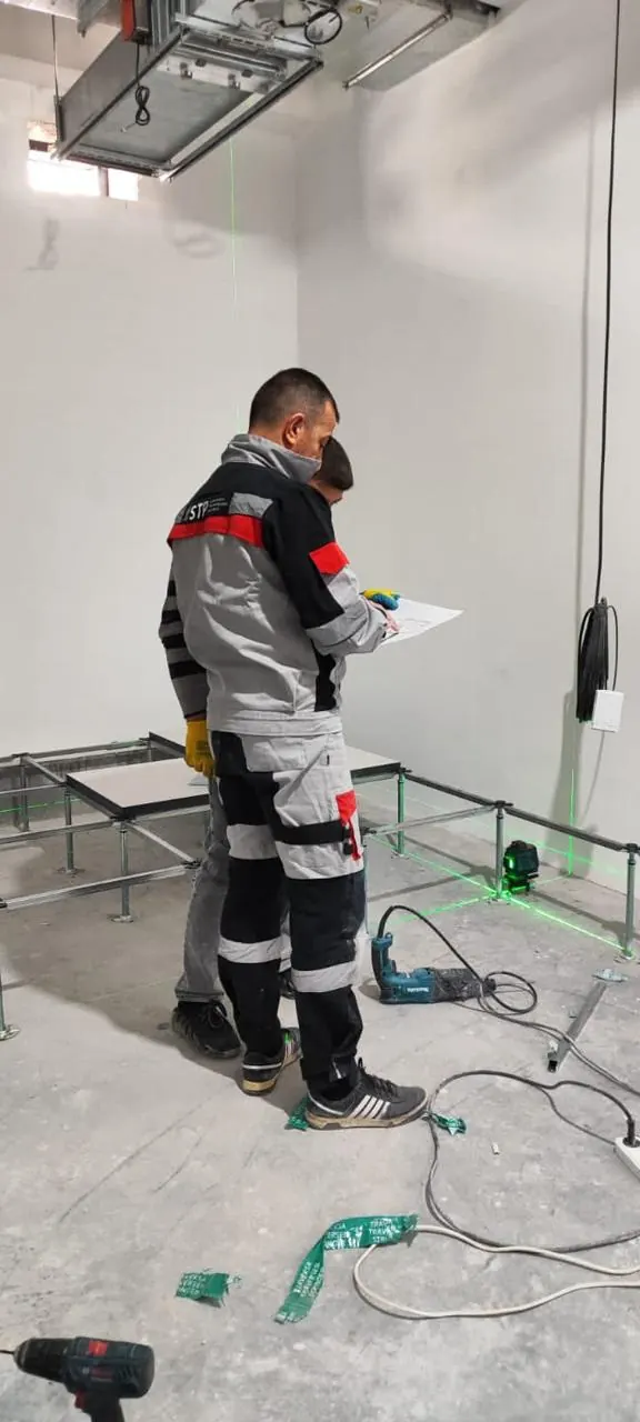 Two STP engineers in branded workwear checking a drawing together while setting out a raised access floor, a green cross-line laser projecting across the slab